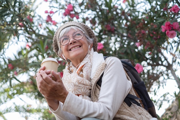 Portrait of senior attractive woman wearing glasses and cap sitting outdoors in public park holding a coffee cup while listening to music by earphones