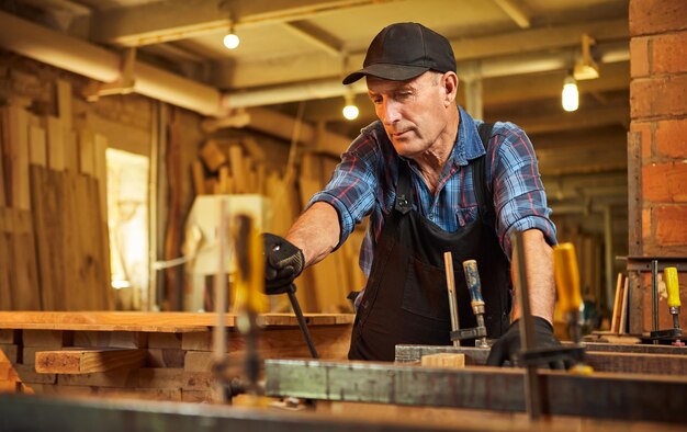 Portrait of a senior carpenter in uniform gluing wooden bars with hand pressures at the carpentry manufacturing