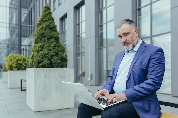 Portrait of senior grayhaired businessman outside office building man working and typing on laptop