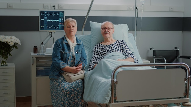 Portrait of senior man in hospital ward bed receiving assistance from wife, waiting on medical advice to cure sickness. Old patient and woman getting ready for consultation at clinic.