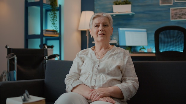 Portrait of senior woman with physical injury sitting on couch. Person looking at camera and sitting next to wheelchair in living room, using mobility equipment for transportation.