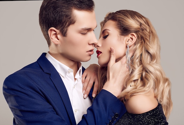 Portrait of sensual beautiful young couple dressed in formal clothes: woman in fashion luxury black dress and man wearing blue classical suit isolated on white wall.