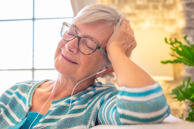 Portrait of serene senior woman wearing earphones listening to music while sitting on sofa at home relaxed elderly lady with closed eyes