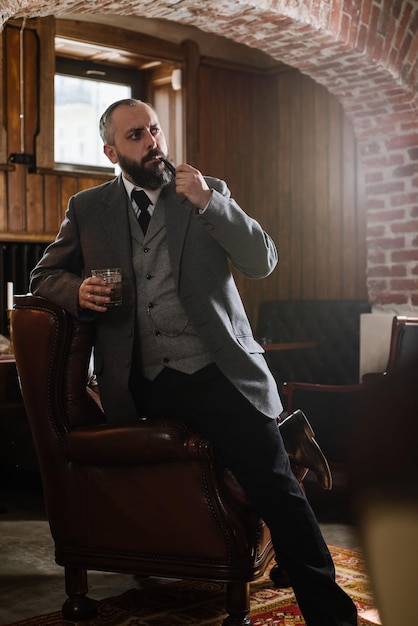 Portrait of serious bearded man with pipe holding glass of whiskey wearing suit and sitting on a big arm chair