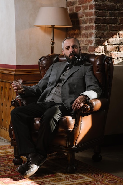 Portrait of serious bearded man with pipe holding glass of whiskey wearing suit and sitting on a big arm chair