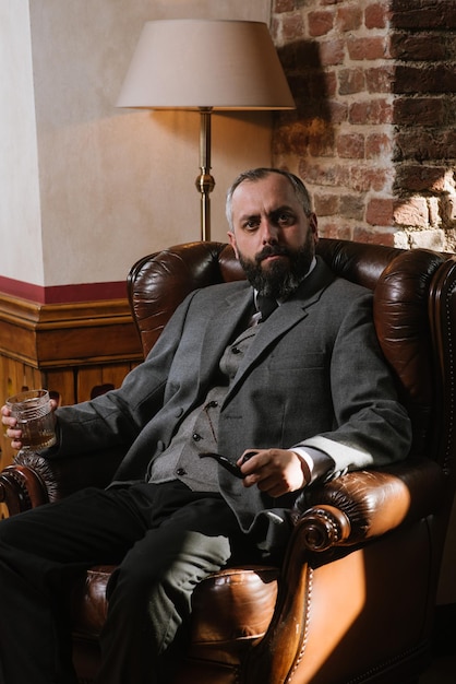 Portrait of serious bearded man with pipe holding glass of whiskey wearing suit and sitting on a big arm chair