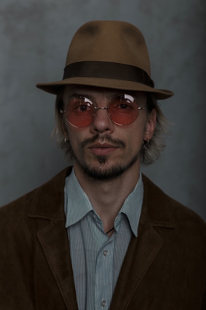 Portrait of a serious bearded young man in vintage red glasses in an elegant hat in a fashionable brown jacket in a classic striped shirt in the studio near a gray wall. brutal guy.