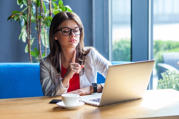 Portrait of serious bossy beautiful stylish brunette young woman in glasses sitting, looking at her laptop screen on video call and warning, giving advice. indoor studio shot, cafe, office background.