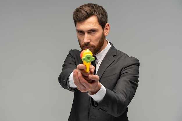 Portrait of a serious businessman playing with water gun against grey background and looking at the camera with calm face