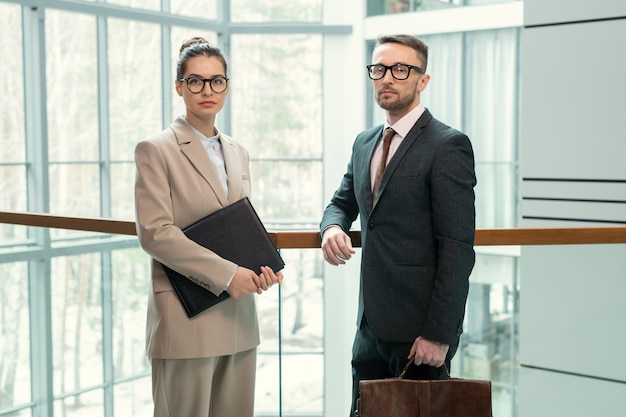 Portrait of serious colleagues in formal wear standing at glassy railing and leaning on it in business hall