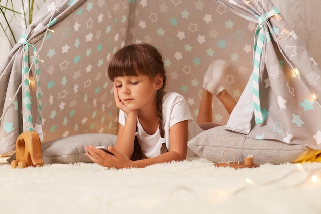 Portrait of serious concentrated girl with dark hair and pigtails wearing white t shirt and leggins lying on the floor and watching cartoons spending time alone in cozy teepee tent