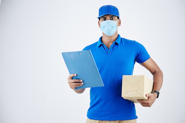 Portrait of serious confident young delivery man in medical mask holding document and package