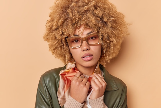 Portrait of serious curly haired woman wears spectacles sas has bad eyesight looks directly at camera dressed in leather jacket poses against brown background. People facial expressions and style