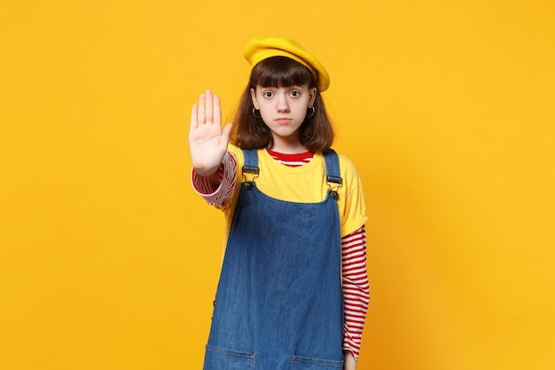 Portrait of serious girl teenager in french beret, denim sundress showing stop gesture with palm isolated on yellow background in studio. People sincere emotions lifestyle concept. Mock up copy space.