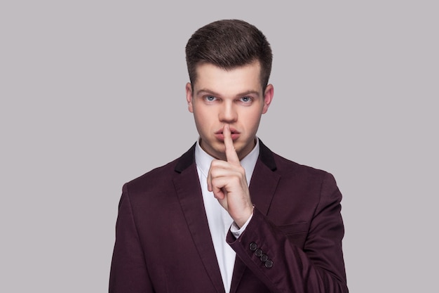 Portrait of serious handsome young man in violet suit and white shirt, standing, looking at camera with silence sign and warning. indoor studio shot, isolated on grey background.