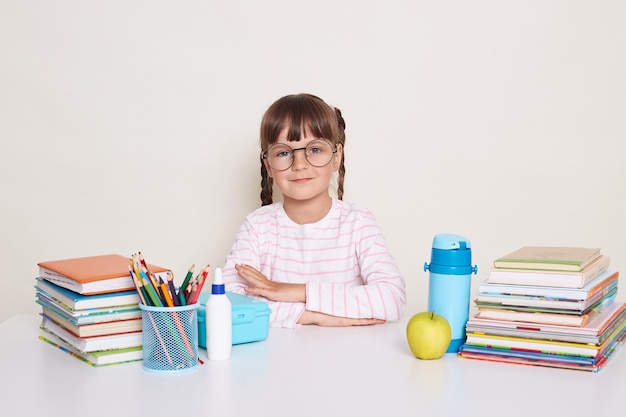 Portrait of serious little schoolgirl with pigtails and glasses wearing striped shirt sitting at the desk surrounded with books and pens keeps hands folded looking at camera waiting for teacher