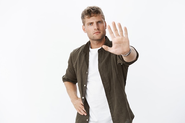 Portrait of serious-looking sexy and masculine serious-looking fair-haired man extending palm in hold motion, prohibiting step further, warning, posing over gray wall.