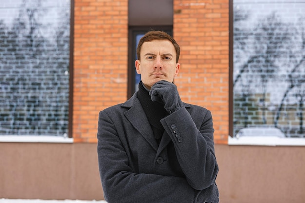 Portrait of serious male entrepreneur in winter coat touching chin and looking at camera on street