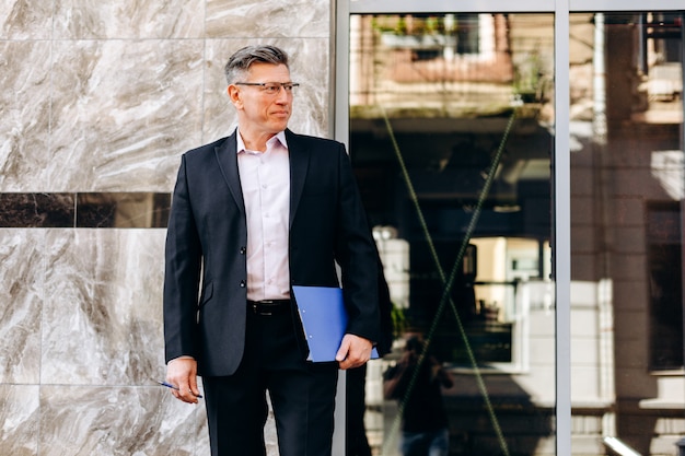 Portrait of serious senior man in suit standing and holding a paper outdoor . 