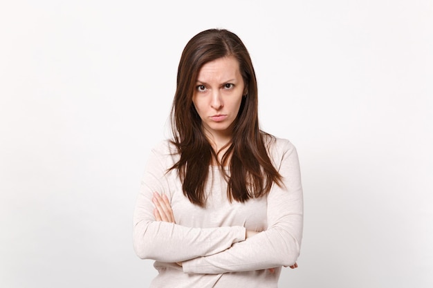 Portrait of serious strict young woman in light clothes looking camera holding hands crossed isolated on white wall background in studio. People sincere emotions lifestyle concept. Mock up copy space.