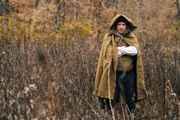 Portrait of serious tourist man wearing green raincoat tent standing in thicket of bushes in cold overcast day and looking at camera