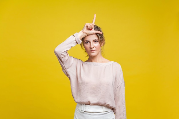 Portrait of serious upset woman with fair hair in casual beige blouse standing showing loser gesture with fingers on her head looking at camera indoor studio shot isolated on yellow background