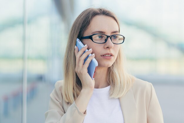 Portrait of a serious woman wearing glasses blonde holding a mobile phone talking on a smartphone near the office