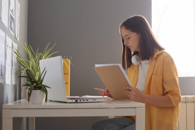 Portrait of serious woman with brown hair sitting at table with laptop analyzing work results young adult accountant doing paperwork at workplace calculating