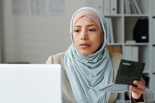 Portrait of serious young adult muslim woman wearing hijab sitting in embassy office using laptop to