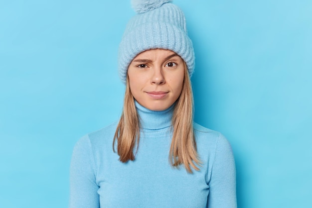 Portrait of serious young woman looks indignant at camera raises eyebrows wears knitted hat and turtleneck listens information attentively isolated over blue background. Human reactions concept