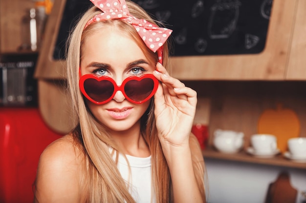 Portrait of sexy blonde long hair girl wearing heartssunglasses looking on the camera in the red loft comfortable sunny kitchen