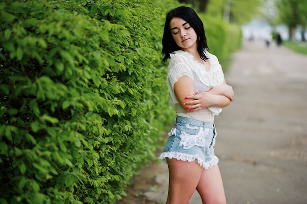 Portrait of sexy brunette girl on women's jeans shorts and white blouse against green spring bushes.