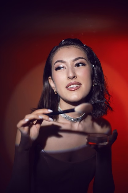 Portrait of a sexy fashionable makeup artist woman in a white skirt with a black powder brush and box stands in the studio red light background