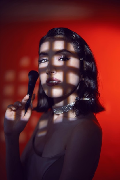 Portrait of a sexy fashionable makeup artist woman in a white skirt with a black powder brush stands in the studio red light with gobo masks cell