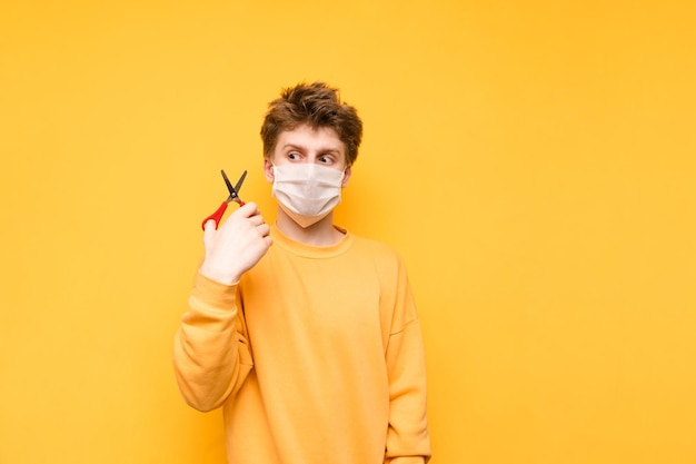 Portrait of a shaggy man in a medical mask on a yellow background holding a scissors for a haircut