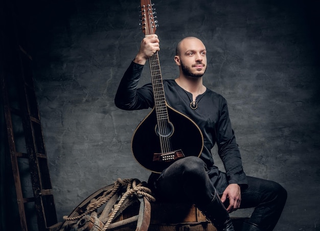 Portrait of a shaved head male musician dressed in old Celtic clothes sits on a wooden box and performing old mandolin.