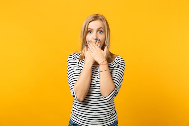 Portrait of shocked amazed young woman in striped clothes covering mouth with hands isolated on yellow orange wall background in studio. People sincere emotions, lifestyle concept. Mock up copy space.