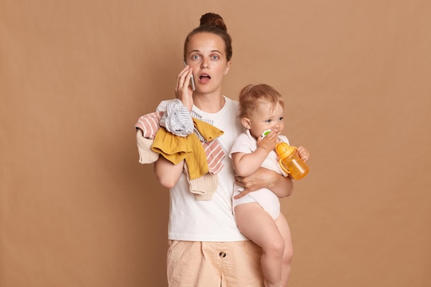 Portrait of shocked beautiful woman with bun hairstyle wearing white t shirt standing with her infant daughter and talking on phone posing with open mouth isolated over brown background