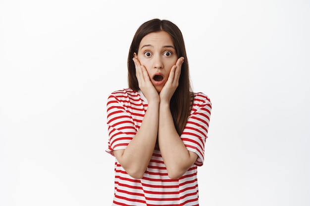 Portrait of shocked brunette woman gasp, drop jaw and look concerned, holding hands on face startled, staring at smth worrying, standing over white background.