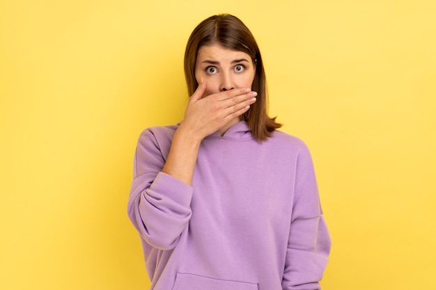 Portrait of shocked dark haired woman put hand on mouth and looking with fear in her eyes keeping terrible secret wearing purple hoodie Indoor studio shot isolated on yellow background