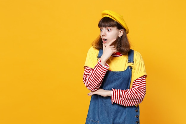 Portrait of shocked girl teenager in french beret, denim sundress looking aside, put hand prop up on chin isolated on yellow background. People sincere emotions, lifestyle concept. Mock up copy space.