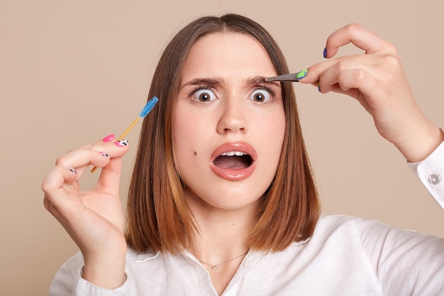 Portrait of shocked scared female in beauty salon holding eyebrow brush and tweezers looking at camera with big eyes being shocked after plucking eyebrow procedure