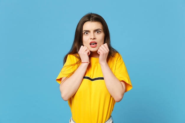 Portrait of shocked scared young woman in vivid casual clothes looking camera, keeping mouth open, clenching fists near face isolated on blue background. People lifestyle concept. Mock up copy space.