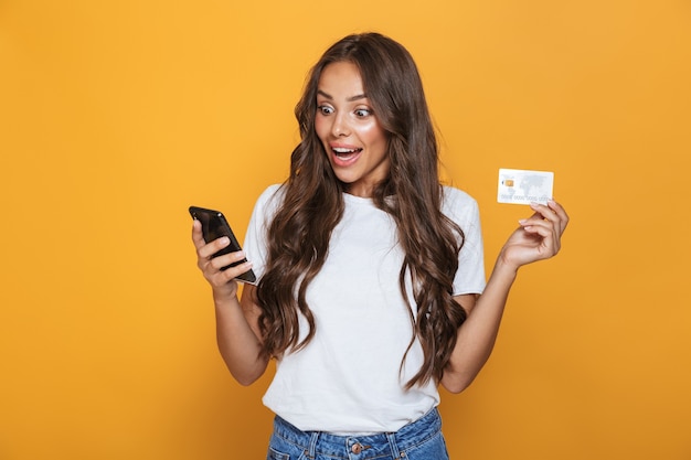 Portrait of a shocked young girl with long brunette hair standing over yellow wall, holding mobile phone, showing plastic credit card