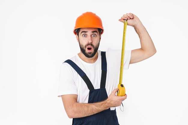 Portrait of shocked young man builder in helmet posing isolated over white wall holding centimeter.