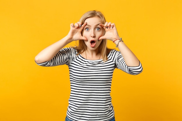 Portrait of shocked young woman in striped clothes keeping mouth wide open, stretching eyelids isolated on yellow orange wall background. People sincere emotions lifestyle concept. Mock up copy space.