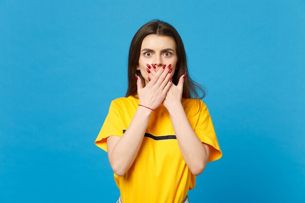 Portrait of shocked young woman in vivid casual clothes looking camera, covering mouth with hands isolated on bright blue wall background in studio. People lifestyle concept. Mock up copy space.