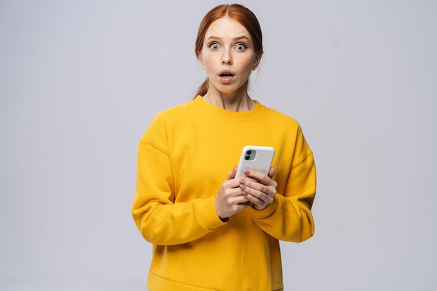 Portrait of shocked young woman wearing yellow sweater holding mobile phone and looking at camera