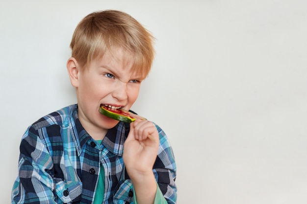 Portrait shot of angry little blond boy with blue eyes biting lollipop over white wall. Pretty little child posing