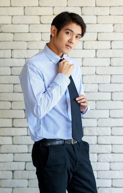 Portrait shot of Asian young happy business office worker wears blue stripe long sleeve shirt standing smiling put hands in pants pockets posing against  white brick wall.